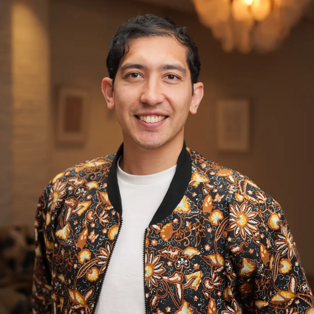 A young man with dark hair and brown eyes, smiling and wearing a white t-shirt with a floral jacket.