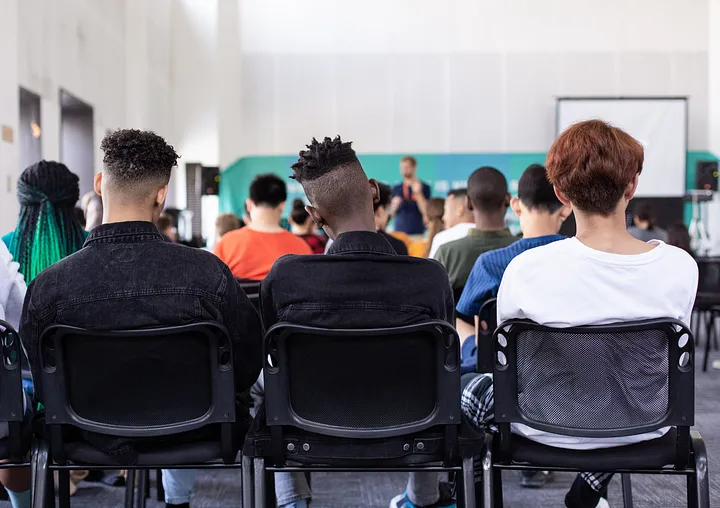 People facing the front of a room whilst sitting on chairs, their backs visible 