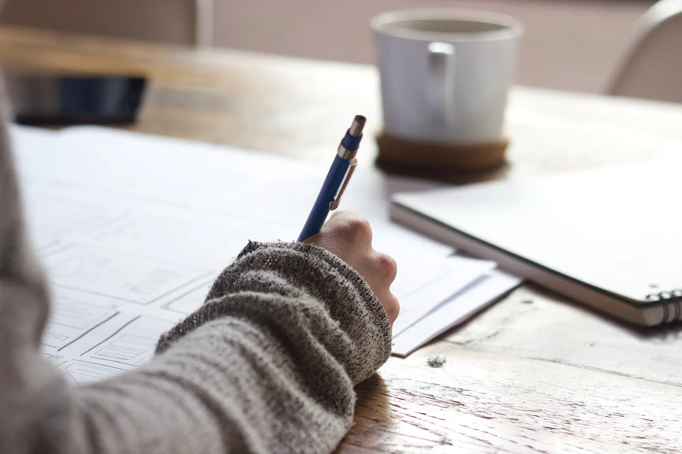A person wearing a grey jumper is writing on a white sheet of paper. There's a mug in the picture at arms length. 